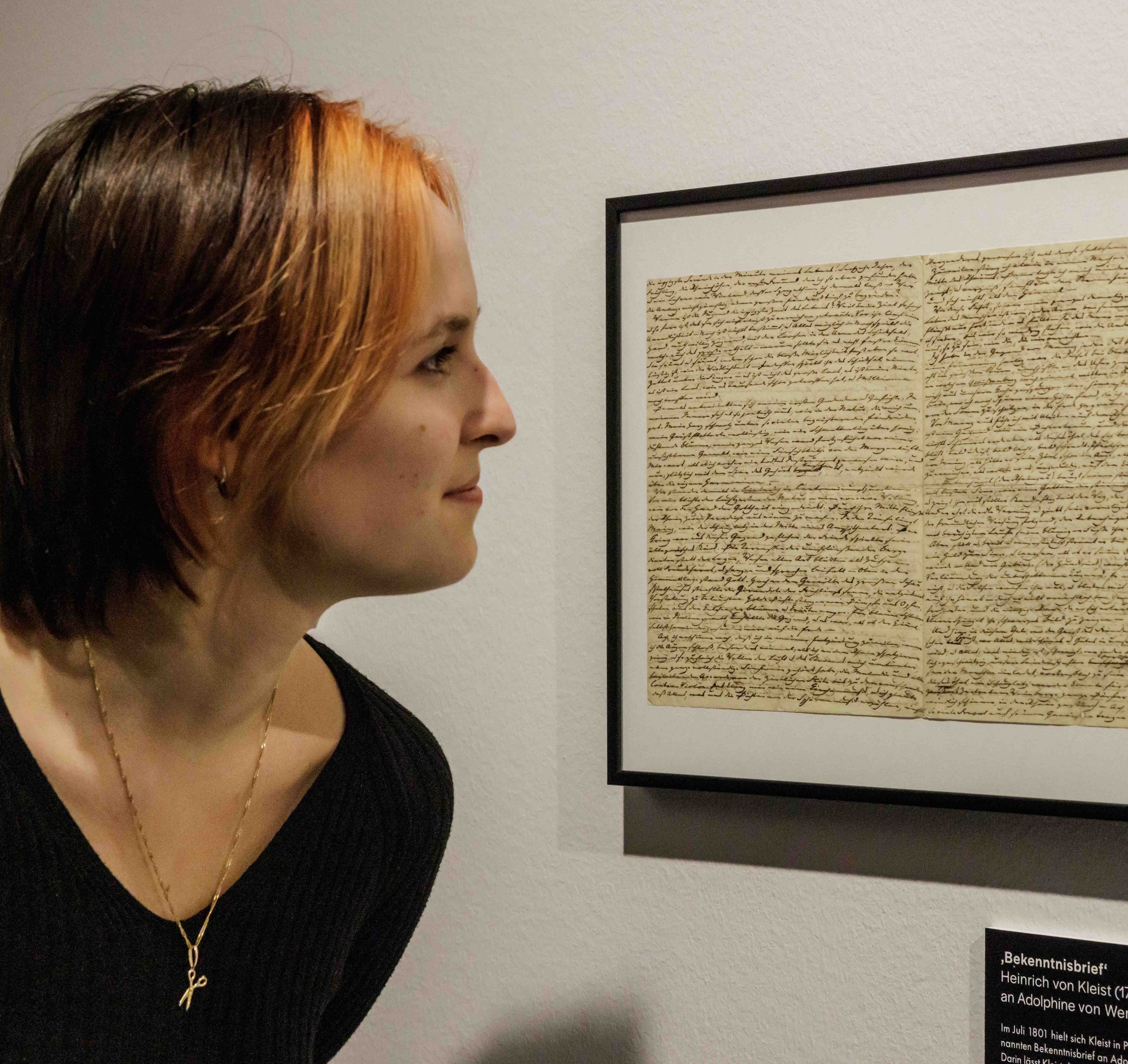 A person with short hair and a black sweater examines a framed, handwritten letter on a wall. The setting is a museum or gallery, conveying curiosity.