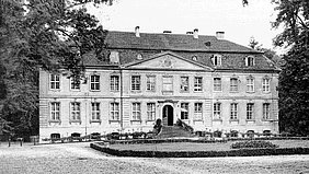 Historisches Herrenhaus umgeben von Bäumen, mit einer eleganten Fassade und einem gepflegten Garten im Vordergrund.