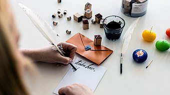 Eine Person schreibt mit einer weißen Feder auf ein Blatt Papier, das auf einem weißen Tisch liegt. Die Handschrift ist kalligrafisch und schwarz. Auf dem Tisch stehen ein brauner Briefumschlag, ein Glas mit schwarzer Tinte, mehrere Holzstempel, ein weiteres Blatt Papier, bunte Wachskerzen in Grün, Gelb und Blau sowie eine brennende Kerze. Die Szene wirkt ruhig und konzentriert.
