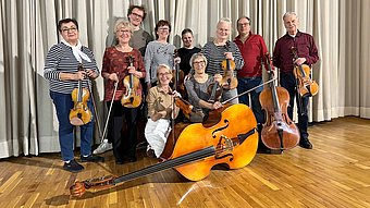 Eine Gruppe von Menschen steht auf einem Parkettboden vor einem hellen Vorhang. Sie halten verschiedene Streichinstrumente, darunter Violinen, Bratschen, Celli und einen Kontrabass, der auf dem Boden liegt. Die Personen sind überwiegend leger gekleidet und blicken in Richtung Kamera.
