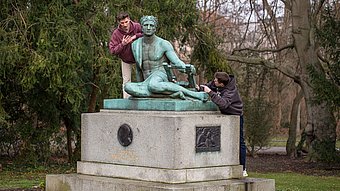 Eine bronzene Statue auf einem steinernen Sockel steht in einer Parkanlage. Die Figur stellt eine sitzende Person mit einer antiken Leier dar. Zwei Personen lehnen sich von beiden Seiten an die Statue, eine links, eine rechts. Beide Personen tragen Freizeitkleidung. Im Hintergrund sind Bäume und Sträucher ohne Laub zu sehen.