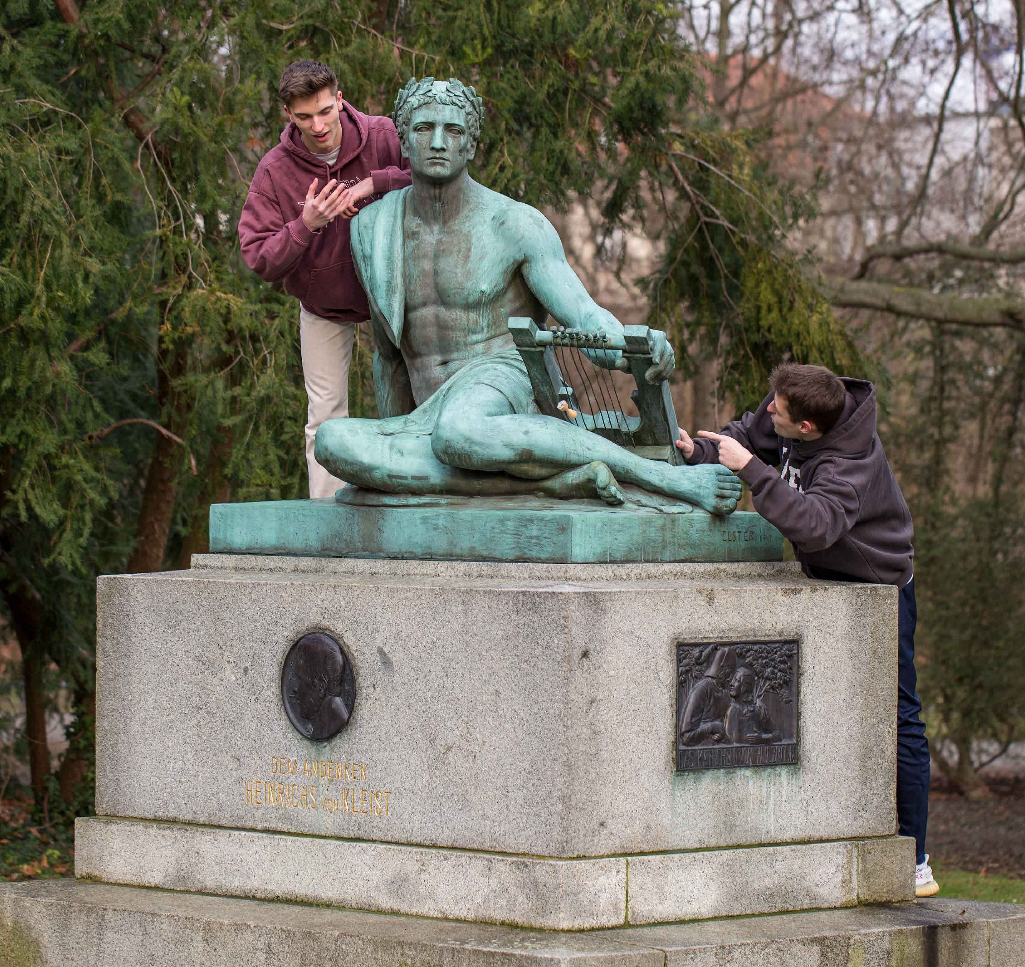 Eine bronzene Statue auf einem steinernen Sockel steht in einer Parkanlage. Die Figur stellt eine sitzende Person mit einer antiken Leier dar. Zwei Personen lehnen sich von beiden Seiten an die Statue, eine links, eine rechts. Beide Personen tragen Freizeitkleidung. Im Hintergrund sind Bäume und Sträucher ohne Laub zu sehen.
