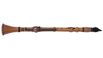 Eine historische Klarinette aus hellem Holz mit mehreren Metallklappen und -ringen liegt waagerecht auf weißem Hintergrund. Das Instrument zeigt eine leicht konische Form mit trichterförmigem Schallbecher links und einem Mundstück mit aufgebundenem Blättchen rechts. Die Oberfläche wirkt glatt, die Mechanik ist schlicht gehalten.