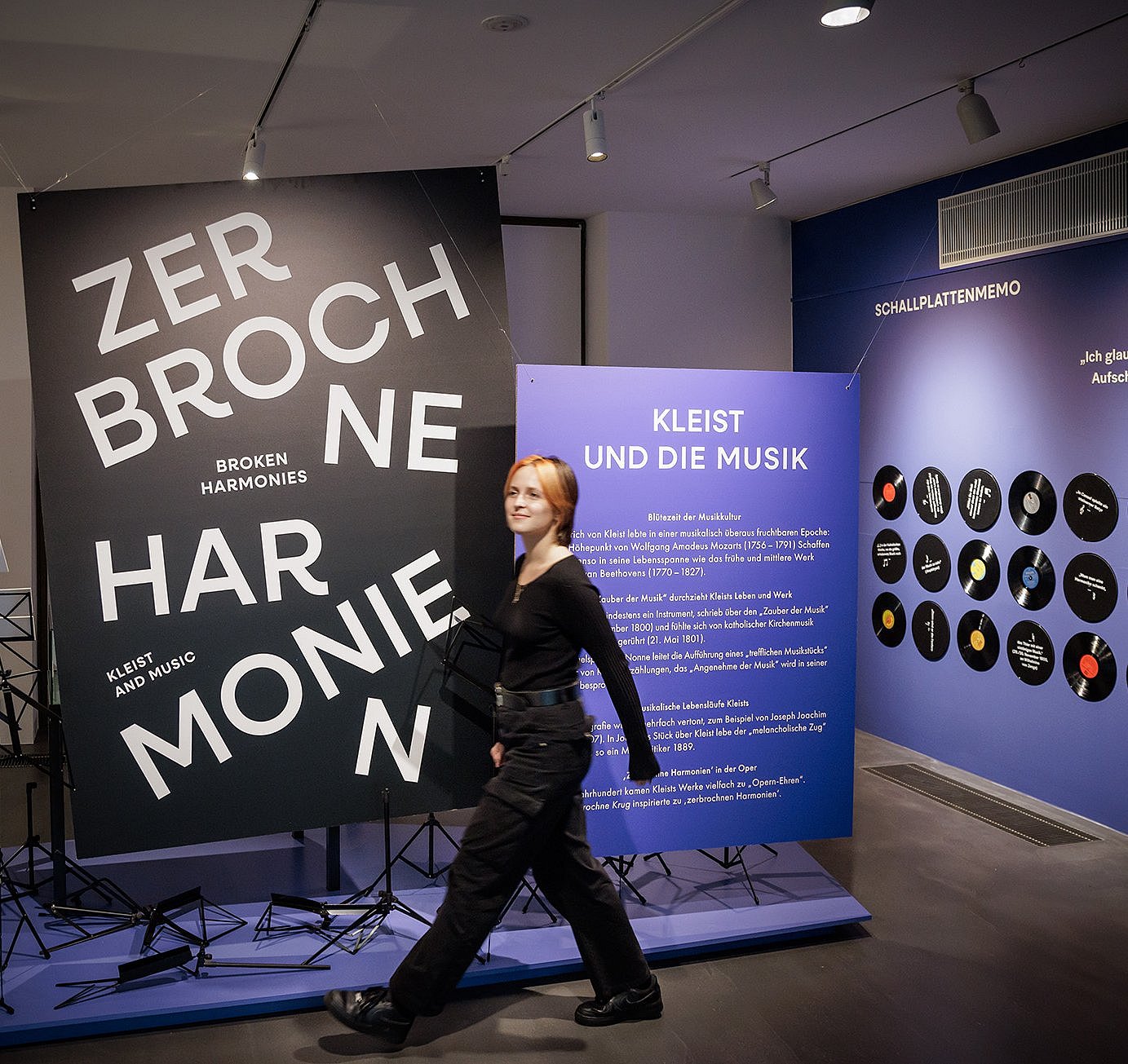 Im Vordergrund steht eine große, schwarze Tafel mit weißer, versetzt angeordneter Schrift: ‚ZERBROCHNE HARMONIEN‘. Links daneben sind Notenständer zu sehen. Im Hintergrund befindet sich eine blaue Wand mit einem Informationstext in weißer Schrift und der Überschrift ‚KLEIST UND DIE MUSIK‘. Rechts davon sind runde, schwarze Tafeln mit grafischen Elementen und kurzen Texten an der Wand angebracht. Der Raum ist hell ausgeleuchtet und wirkt modern gestaltet.