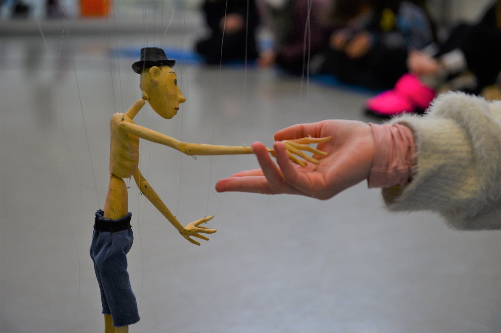 Eine Marionette mit nacktem Oberkörper und Hut, die mit ihrer kleinen Marionettenhand einem Menschen die Hand gibt (nur die Hand des Menschen ist zu sehen)