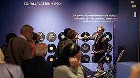 Ein Mann im Anzug steht vor einer blauen Wand mit Schallplatten und spricht zu einer Gruppe von Menschen. Die Schallplatten sind in einer Reihe an der Wand angebracht. Über den Schallplatten steht in großen weißen Buchstaben ein Zitat von Heinrich von Kleist. Die Zuhörerinnen und Zuhörer stehen dicht beieinander und blicken aufmerksam auf den Mann, der mit einer ausladenden Geste spricht. Die Szene spielt in einem Ausstellungsraum mit gedämpfter Beleuchtung.