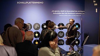Ein Mann im Anzug steht vor einer blauen Wand mit Schallplatten und spricht zu einer Gruppe von Menschen. Die Schallplatten sind in einer Reihe an der Wand angebracht. Über den Schallplatten steht in großen weißen Buchstaben ein Zitat von Heinrich von Kleist. Die Zuhörerinnen und Zuhörer stehen dicht beieinander und blicken aufmerksam auf den Mann, der mit einer ausladenden Geste spricht. Die Szene spielt in einem Ausstellungsraum mit gedämpfter Beleuchtung.