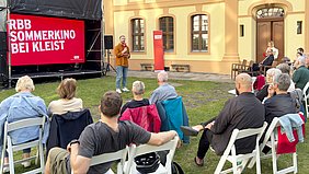 Im Innenhof eines historischen Gebäudes sitzen Menschen auf weißen Klappstühlen und blicken auf eine große Leinwand mit rotem Hintergrund. Darauf steht in weißen Großbuchstaben: ‚RBB SOMMERKINO BEI KLEIST‘. Ein Mann mit Mikrofon steht vor der Leinwand und spricht zum Publikum. Im Hintergrund sind gelb gestrichene Fassaden mit großen Fenstern und ein Türbogen zu sehen. Die Szene spielt bei Tageslicht auf einer Rasenfläche.