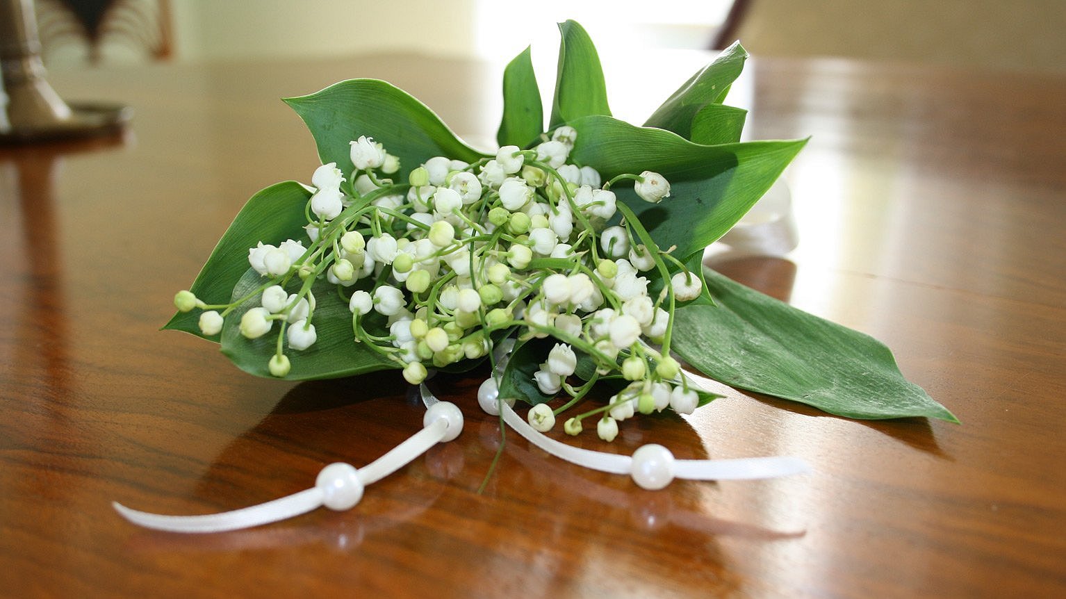 Kleiner Blumenstrauß aus Maiglöckchen mit weißen Perlen und Bändern verziert, der auf einer braunen, versiegelten Tischoberfläche liegt