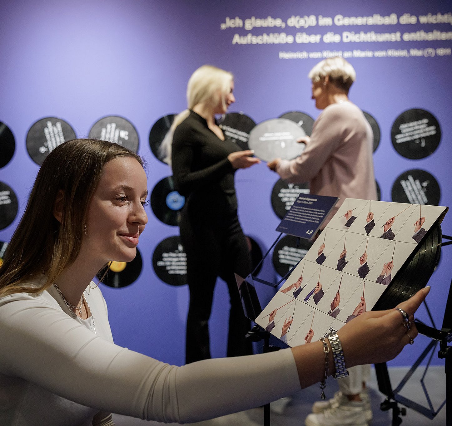 A look at the special exhibition ©Gordon Welters Against a blue wall decorated with vinyl record motifs and a white quote, a person in the foreground holds a square board with a graphic design depicting hands in various gestures. In the background, there are two people, one of whom is holding a vinyl record. The scene takes place at a music or art exhibition.