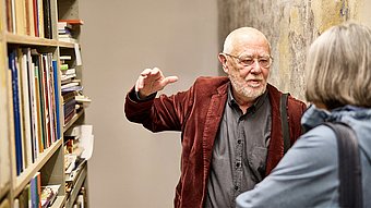 Ein Mann mit Glatze und Brille trägt ein weinrotes Sakko und ein graues Hemd. Er steht in einem schmalen Gang zwischen einem Bücherregal und einer Wand mit abblätternder Farbe. Seine rechte Hand ist erhoben, als würde er etwas erklären. Im Vordergrund ist eine Person mit grauem, kinnlangem Haar und blauer Jacke von hinten zu sehen.