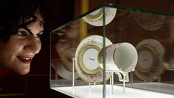 Eine Person mit lockigem, dunklem Haar betrachtet eine Tasse mit Untertasse, die in einer beleuchteten Glasvitrine ausgestellt ist. Die Tasse und die Untertasse sind mit goldfarbenen Rändern und floralen Mustern verziert. Auf der Tasse steht in dunkler Schrift ‚uns‘. Die Szene ist in warmes Licht getaucht.