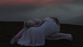 Zwei Personen mit langen, hellen Haaren und weißen, fließenden Gewändern sitzen eng umschlungen am Ufer eines ruhigen Sees. Die Wasseroberfläche spiegelt einen farbenprächtigen Sonnenuntergang mit rosa, orange und violett getönten Wolken wider.