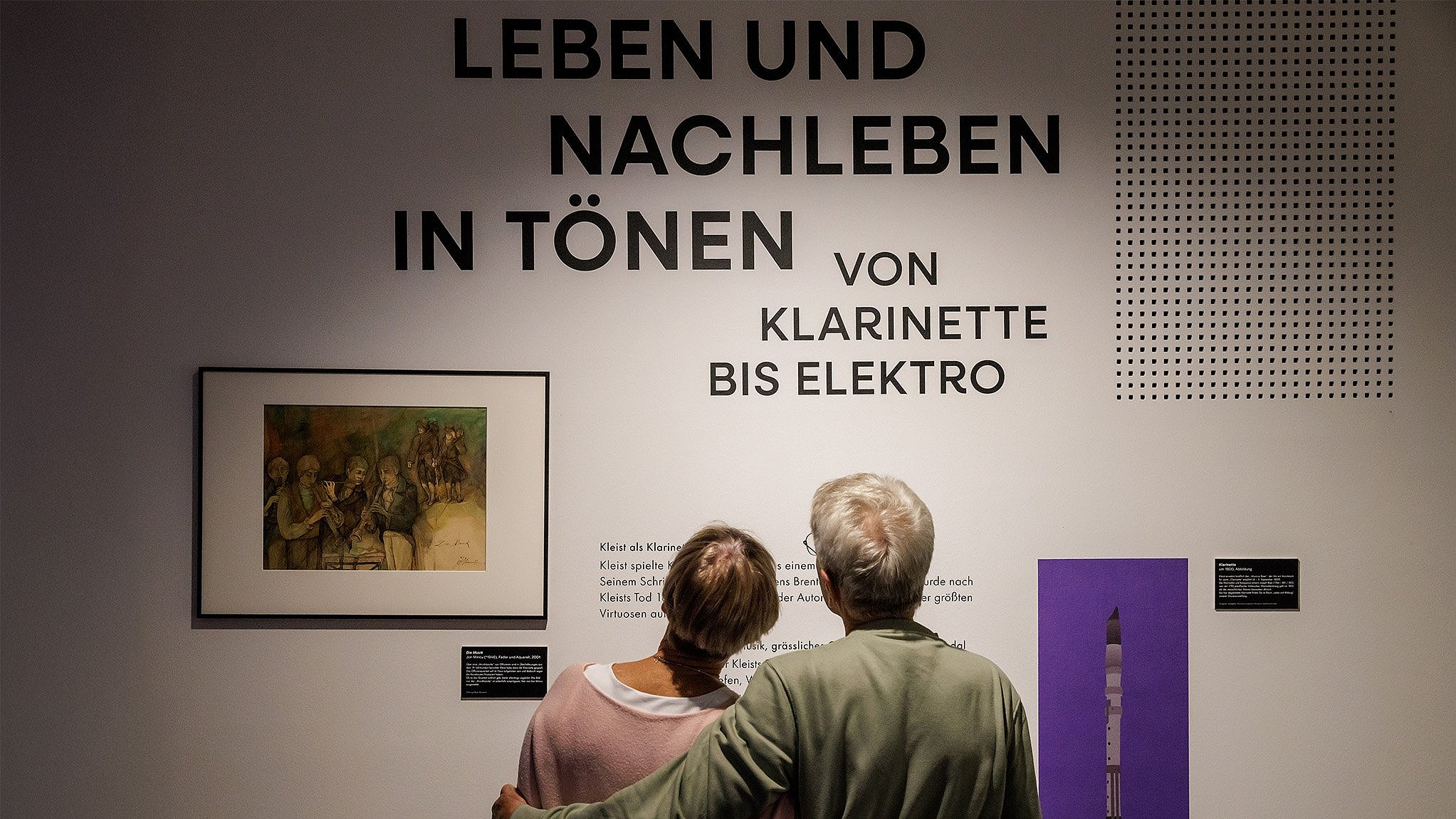 Zwei Personen von hinten umarmt vor einer Wand mit der Aufschrift ‚LEBEN UND NACHLEBEN IN TÖNEN VON KLARINETTE BIS ELEKTRO‘ und einem Bild sowie einer Abbildung einer Klarinette