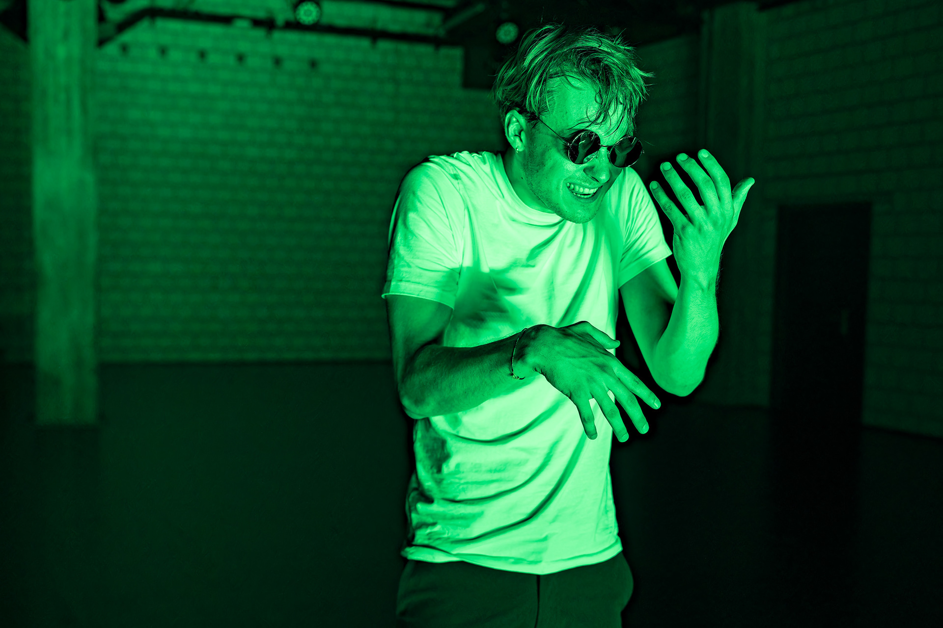 Der Schauspieler Jonas Dumke in einem kargen Raum, in grünes Licht getaucht; der Schauspieler hat wirres Haar, eine schwarze Sonnenbrille und wirkt in seiner Körperhaltung außer sich
